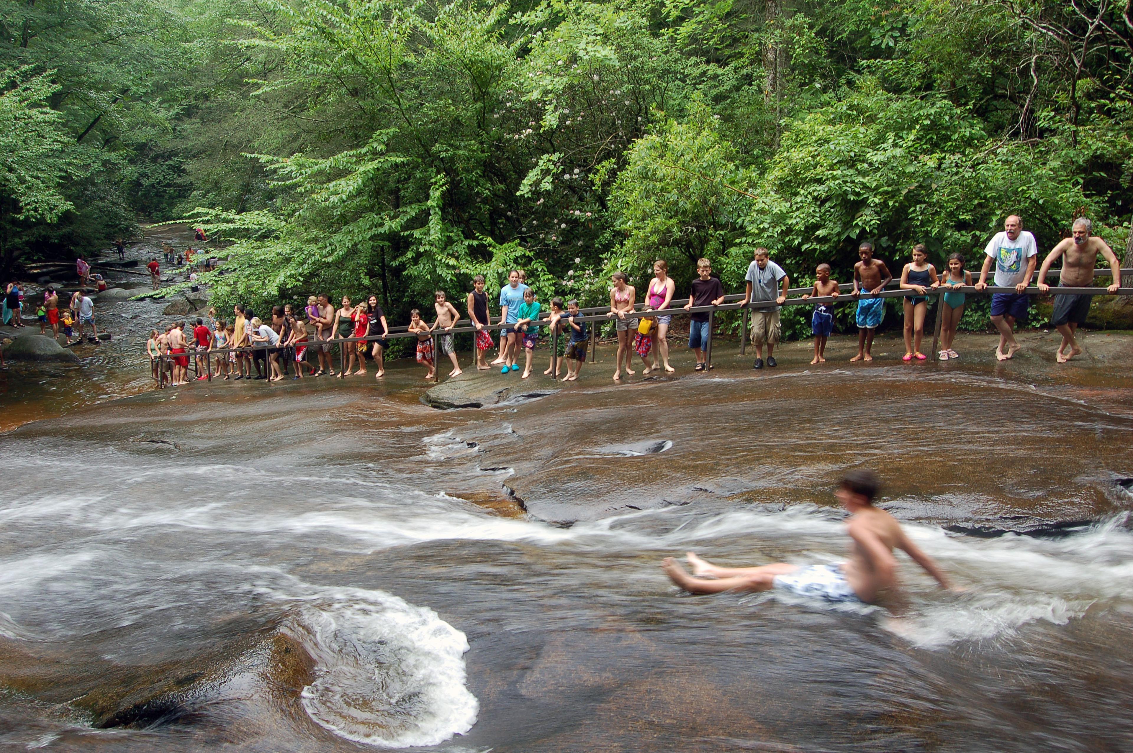 A person slides down the natural waterfall in front of a line of people with green trees in the background