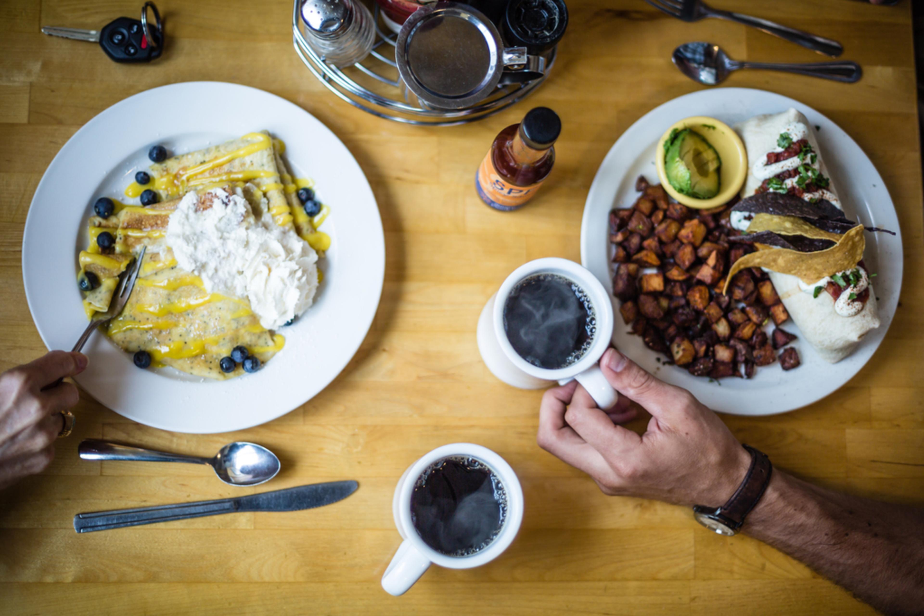 A plate of crepes with whipped cream, a breakfast burrito and two cups of coffee are on a table at an Ashville breakfast restaurant